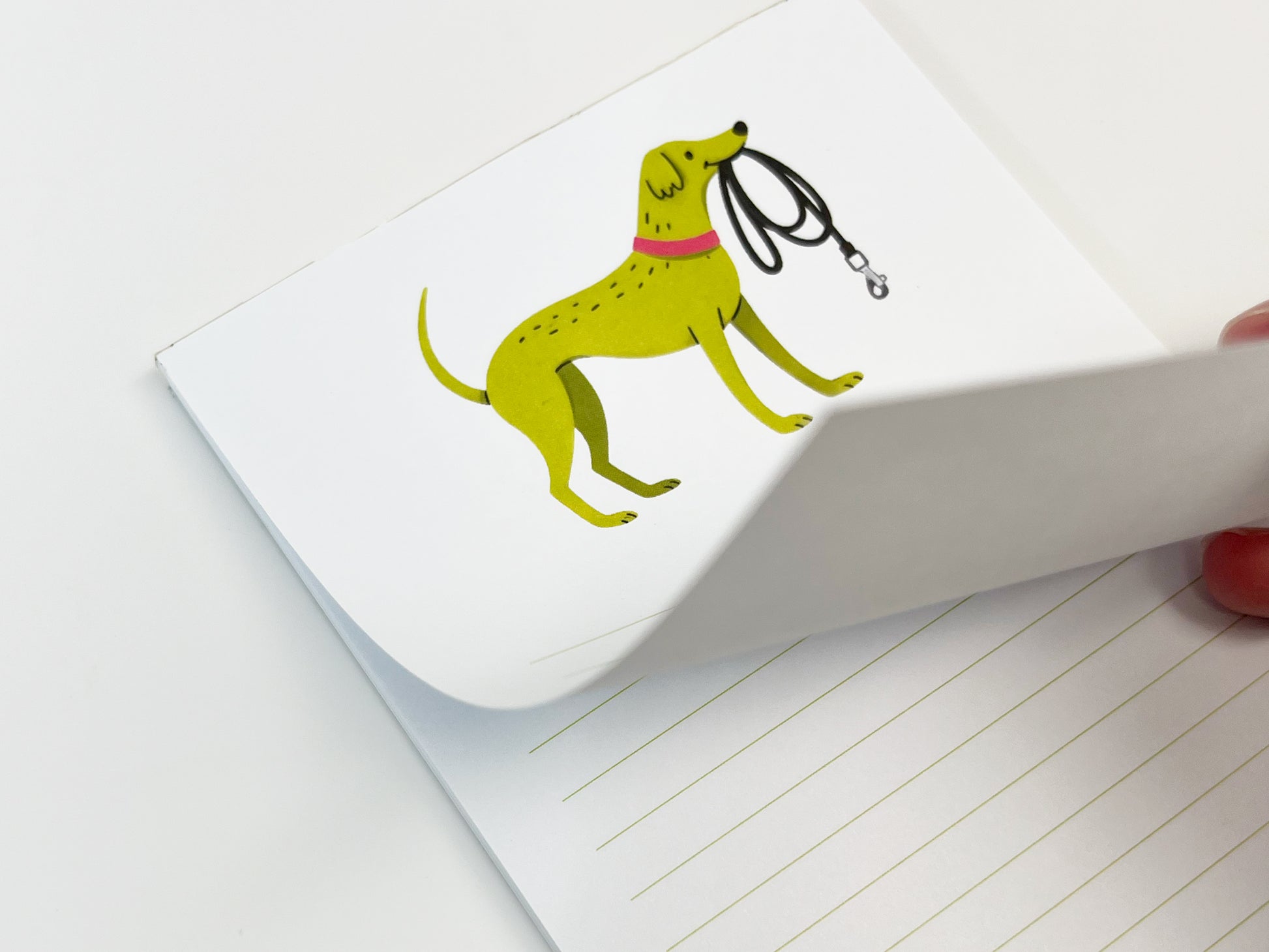 A close-up of a small 4.25 x 5.5 inch lined notepad with the top page partially lifted, showing the thickness of the paper stack. The notepad features an illustrated green dog wearing a pink collar and holding a black leash on the top page, with light green horizontal writing lines visible on the page beneath against a white background.