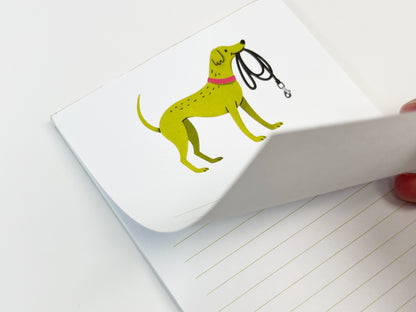 A close-up of a small 4.25 x 5.5 inch lined notepad with the top page partially lifted, showing the thickness of the paper stack. The notepad features an illustrated green dog wearing a pink collar and holding a black leash on the top page, with light green horizontal writing lines visible on the page beneath against a white background.