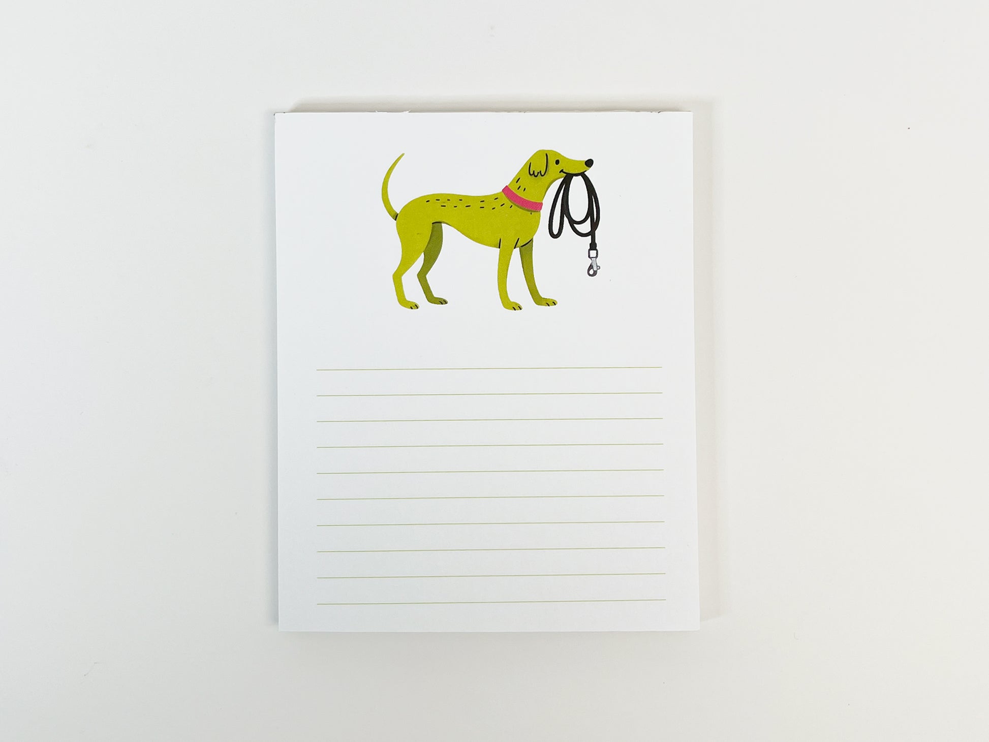 A small 4.25 x 5.5 inch lined notepad photographed flat against a white background. The notepad features an illustrated green dog wearing a pink collar and holding a black leash at the top, with light green horizontal writing lines filling the lower portion of the page.