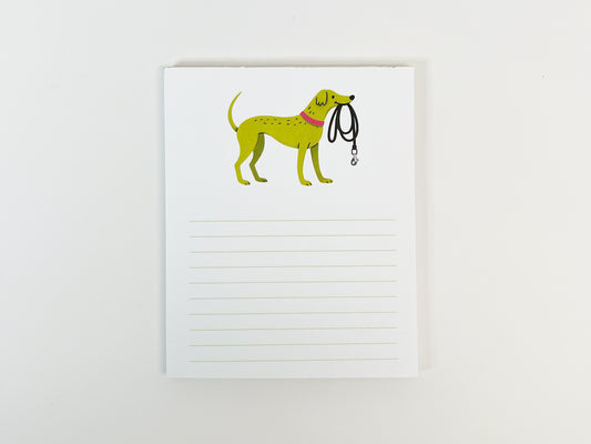 A small 4.25 x 5.5 inch lined notepad photographed flat against a white background. The notepad features an illustrated green dog wearing a pink collar and holding a black leash at the top, with light green horizontal writing lines filling the lower portion of the page.
