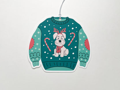 Festive Christmas sweater-shaped gift tag. The sweater is green with playful holiday details, including a cute illustration of a dog wearing reindeer antlers and a red scarf. The sweater is decorated with candy canes and stitched patches on the sleeves, along with snowflakes scattered across the background. A white string is attached to the top for easy attachment to gifts.