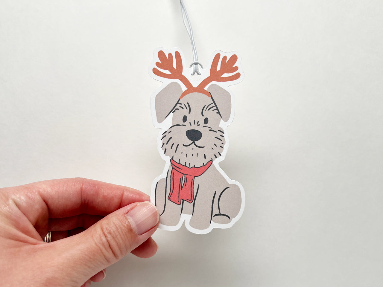A close-up image of a hand holding a festive reindeer-themed Schnauzer gift tag. The handmade gift tag features an adorable gray Schnauzer wearing reindeer antlers and a red scarf, perfect for Christmas gift wrapping and dog lover holiday decor.
