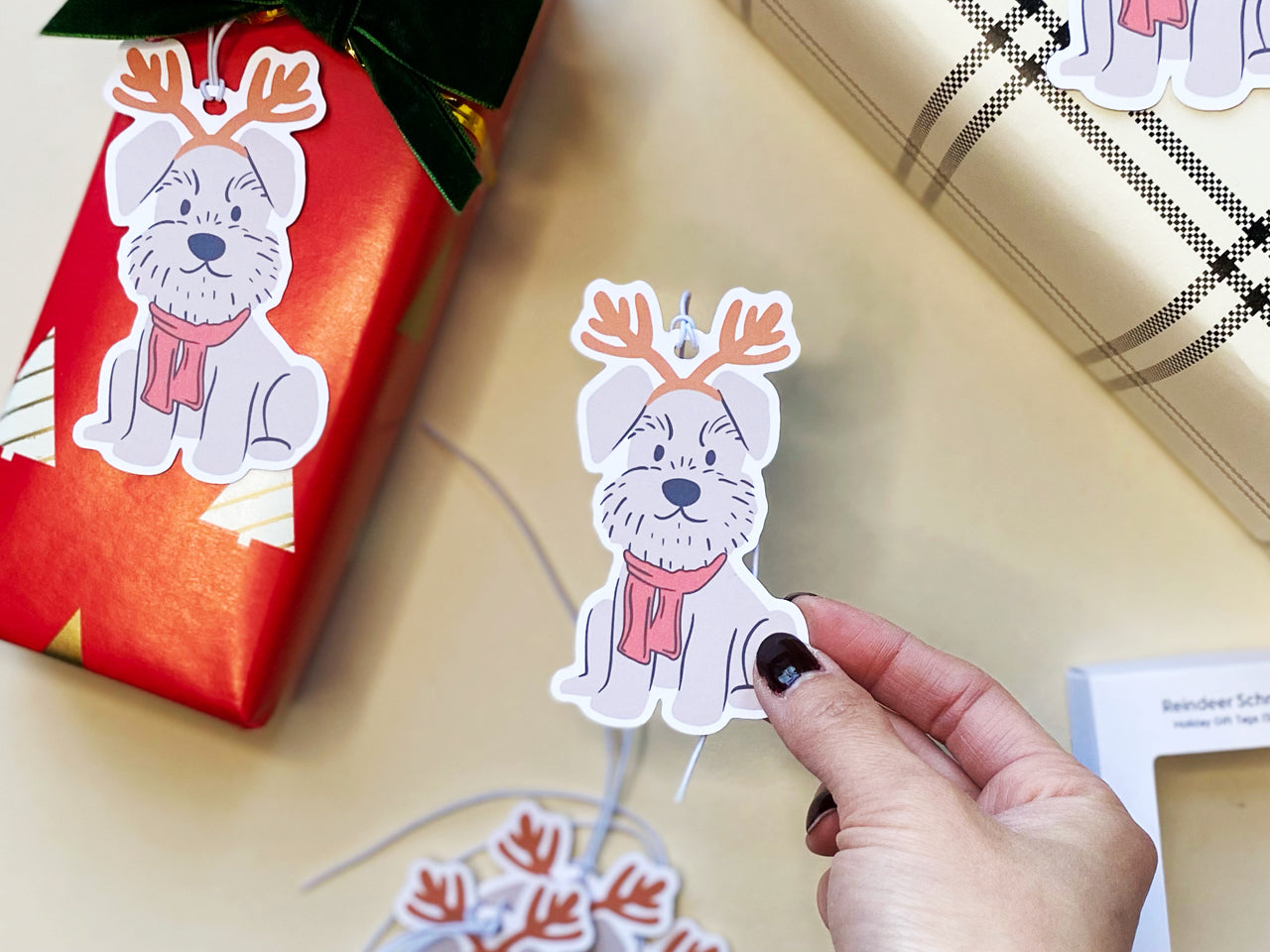 A festive reindeer Schnauzer holiday gift tag held in a hand, surrounded by additional tags and a wrapped red Christmas gift. The tag features a gray Schnauzer with reindeer antlers and a red scarf, pre-attached with an elastic string for easy use.
