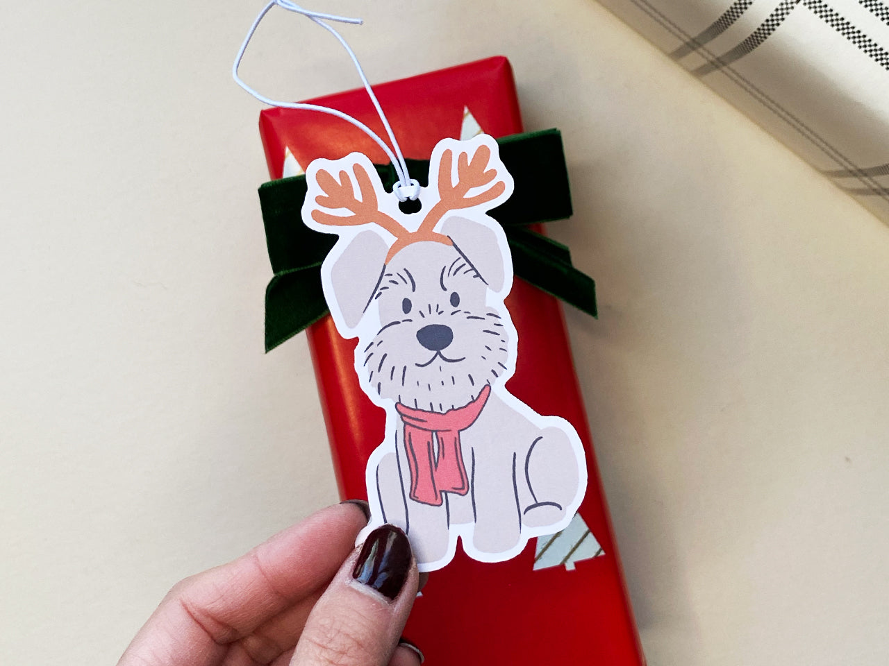A festive reindeer Schnauzer holiday gift tag attached to a beautifully wrapped red Christmas gift with a green velvet bow. The tag features a cute Schnauzer dog with reindeer antlers and a red scarf.