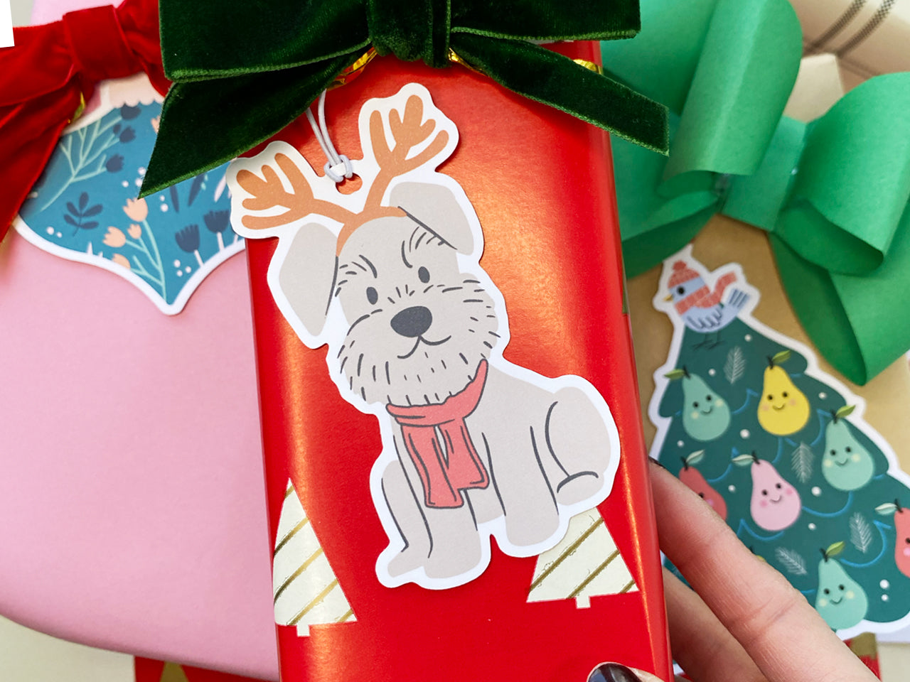 A festive reindeer Schnauzer holiday gift tag attached to a beautifully wrapped red Christmas gift with a green velvet bow. The die cut gift tag features a playful gray Schnauzer with reindeer antlers and a red scarf.