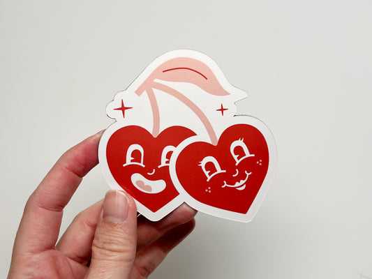 Hand holding a handmade die-cut magnet shaped like two retro cherry hearts with smiling cartoon faces and pink stems. The bold red design makes this customizable wedding favor a fun and unique keepsake for guests to display on fridges or metal surfaces.