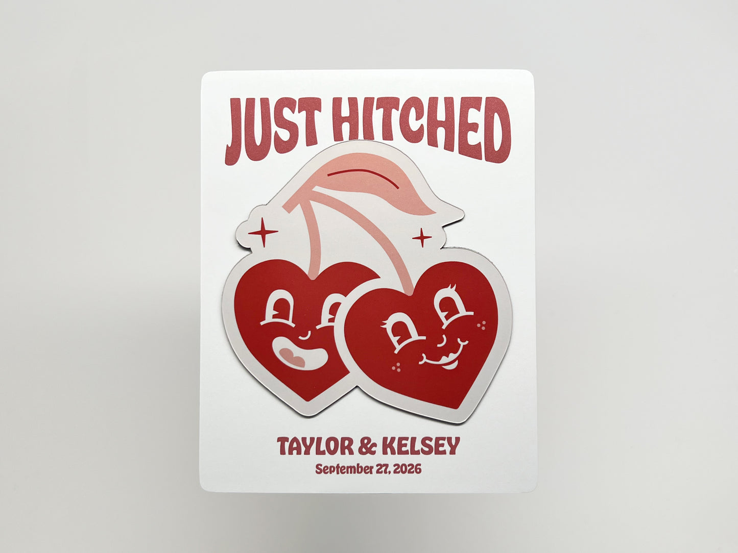 Customizable wedding favor card with a handmade die-cut magnet of two smiling retro cherry hearts. The backing card reads “Just Hitched” with space for personalized names and wedding date, making it a playful keepsake for guests to remember the celebration.