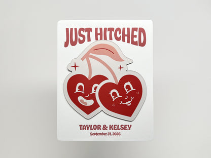 Customizable wedding favor card with a handmade die-cut magnet of two smiling retro cherry hearts. The backing card reads “Just Hitched” with space for personalized names and wedding date, making it a playful keepsake for guests to remember the celebration.
