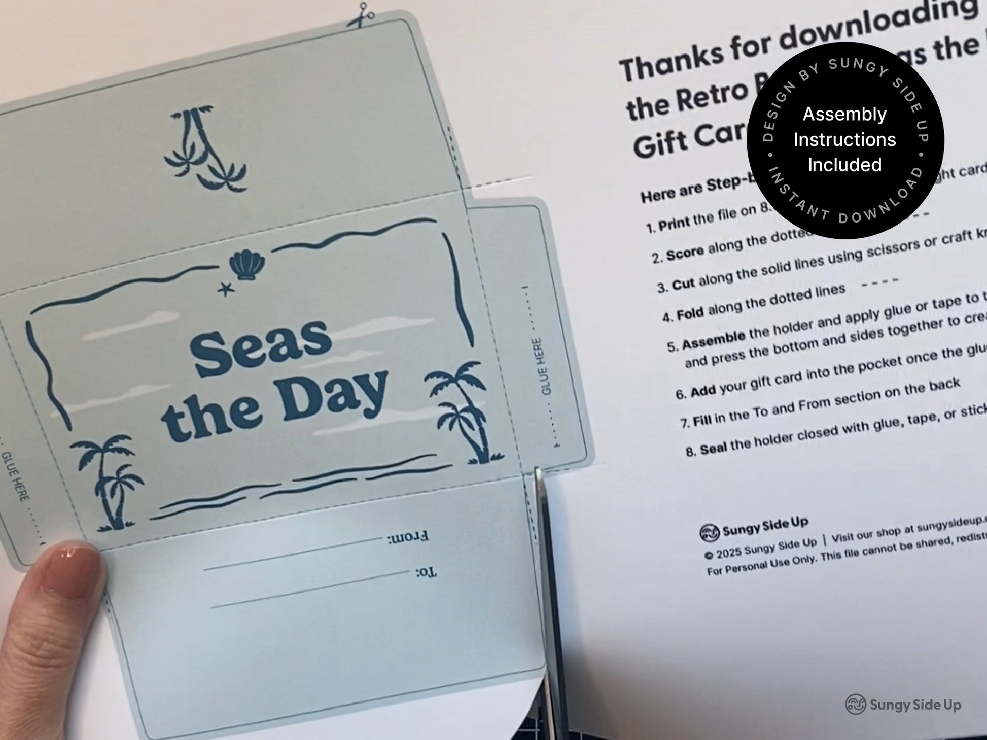 A close-up view shows a person assembling a printable “Seas the Day” gift card holder with a retro beach theme. The unfolded template is printed on light blue paper and includes folding lines, glue tabs, and a “To/From” section. An instruction sheet with step-by-step directions is placed beside it. This DIY project is part of a digital download bundle that includes printable gift card sleeves for summer events, thank you gifts, beach-themed parties, and graduations. Instructions included.