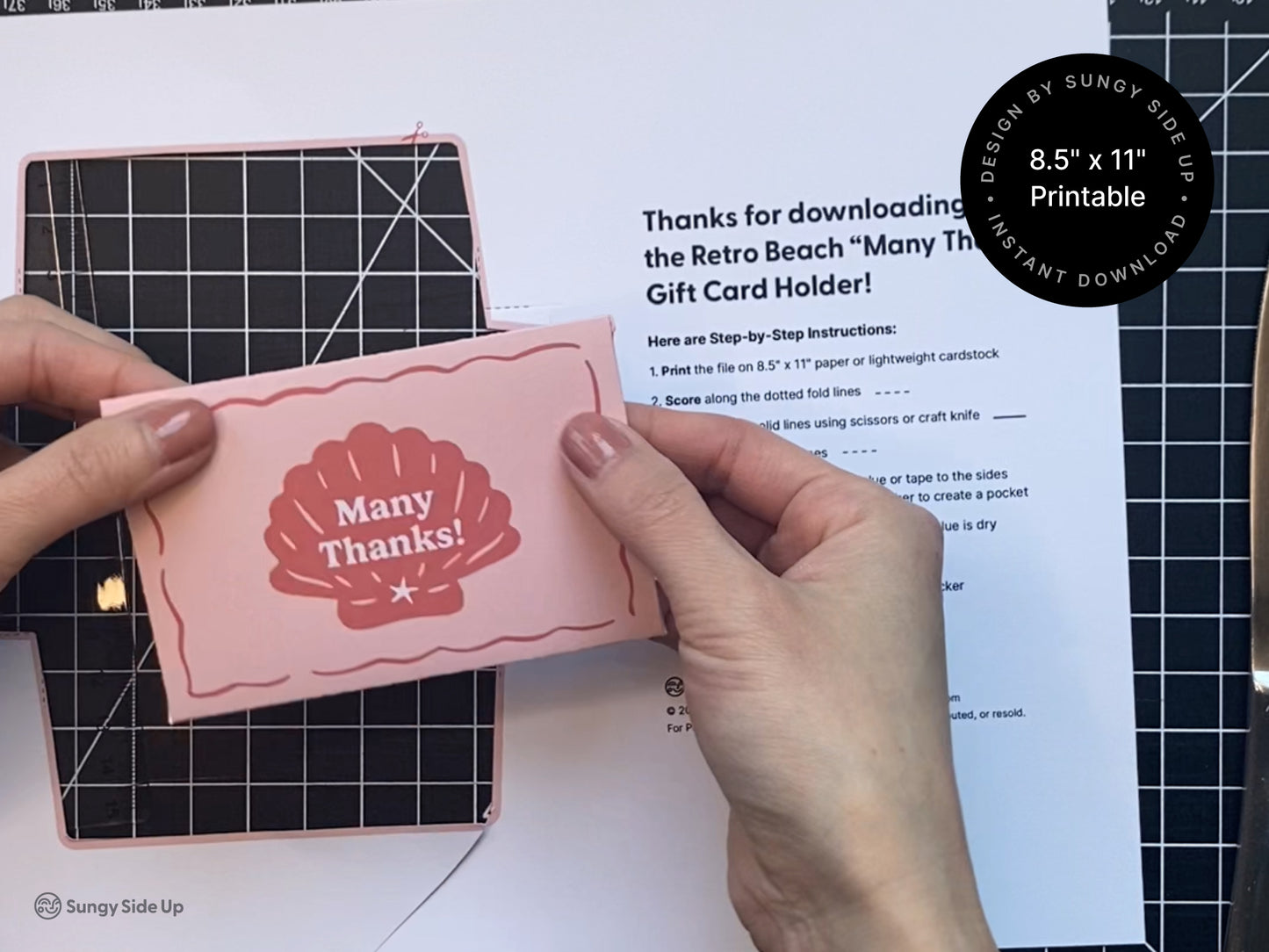 Hands holding a pink and red printable retro-style gift card holder featuring a seashell design with a crafting mat and printed instructions in the background. The printed template and cutting tools are partially visible, indicating an in-progress DIY assembly. The project uses 8.5" x 11" paper or cardstock and includes step-by-step directions. This image showcases the crafting process for a beach-themed thank you gift card envelope, designed for instant download and home printing.
