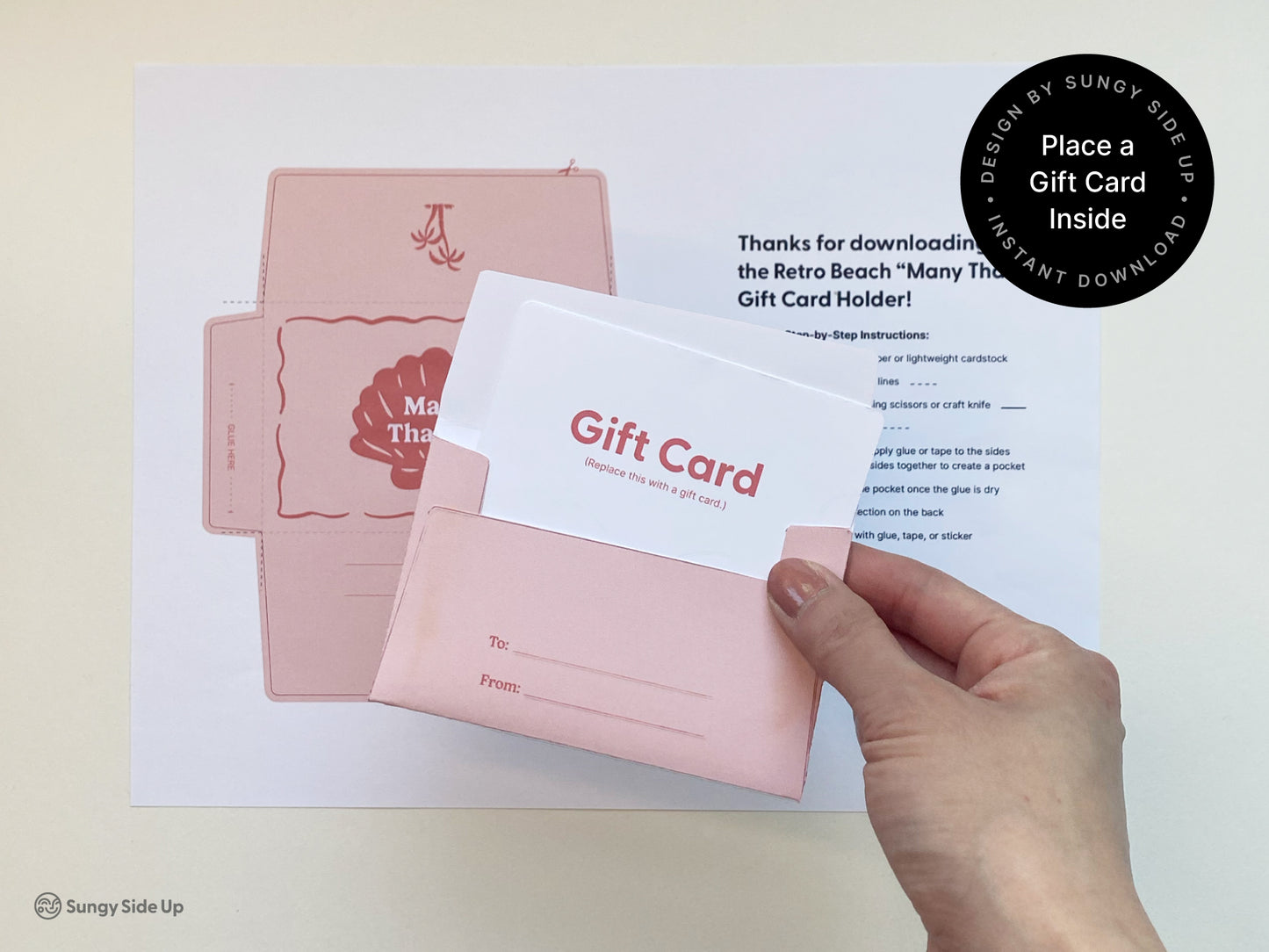 Hand holding a completed printable retro-style gift card holder in pink with a mock gift card inserted inside. The back of the holder includes “To” and “From” lines for personalization. In the background, a printed template and step-by-step instructions are visible, laid out on a flat surface. The scene shows how to use the holder for gifting, ideal for thank you gifts, birthdays, or special occasions. Designed by Sungy Side Up, this item is an instant download for 8.5" x 11" paper.