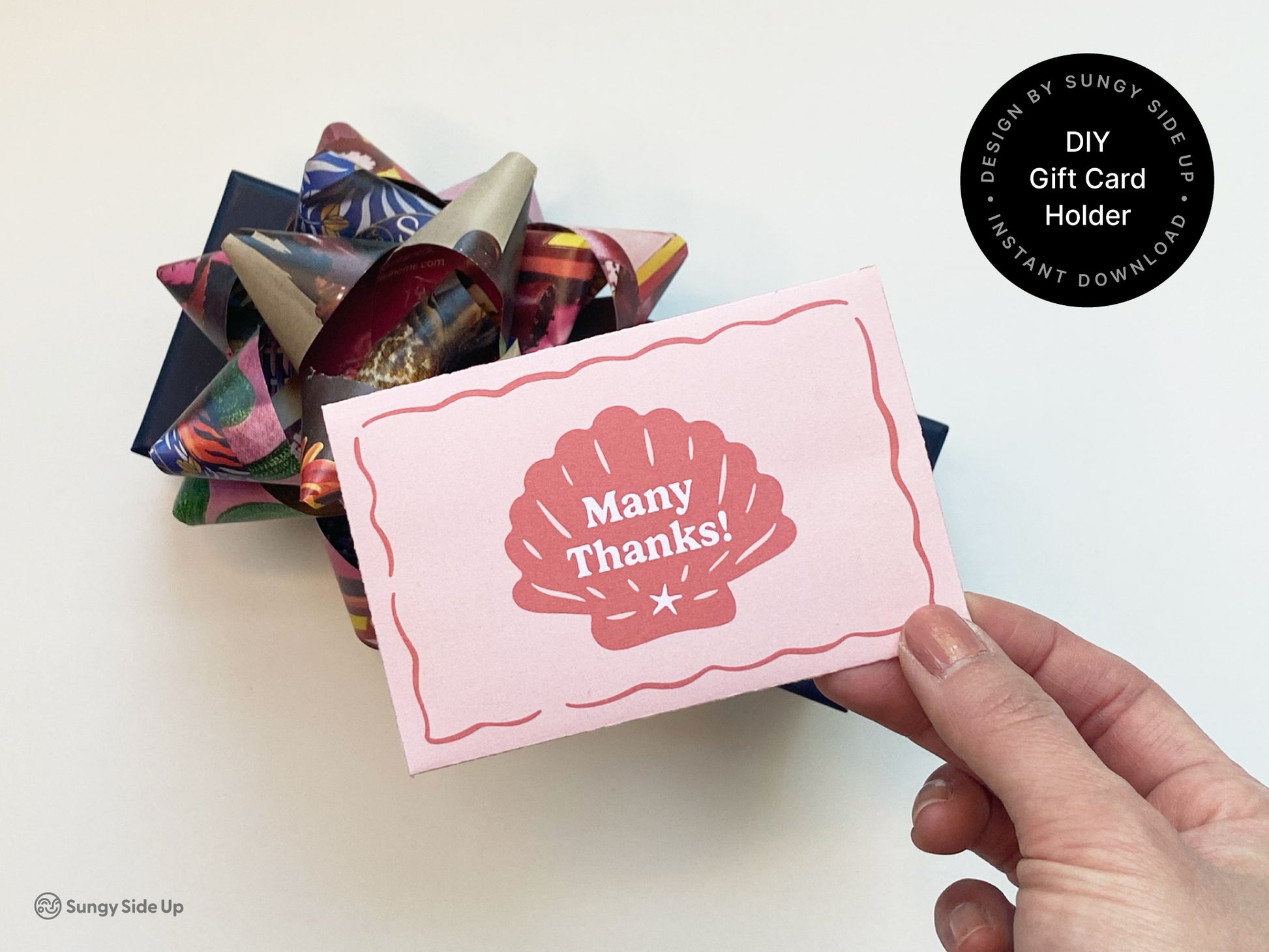 Hand holding a retro-style printable gift card holder in pink and red with a seashell design and scalloped border. In the background, a dark blue gift box is topped with a colorful, multi-patterned bow, suggesting a festive occasion. The scene showcases the DIY gift card holder as a thoughtful addition to a thank-you gift or celebration. This printable design is ideal for birthdays, holidays, or beach-themed events and is available as an instant digital download from Sungy Side Up.