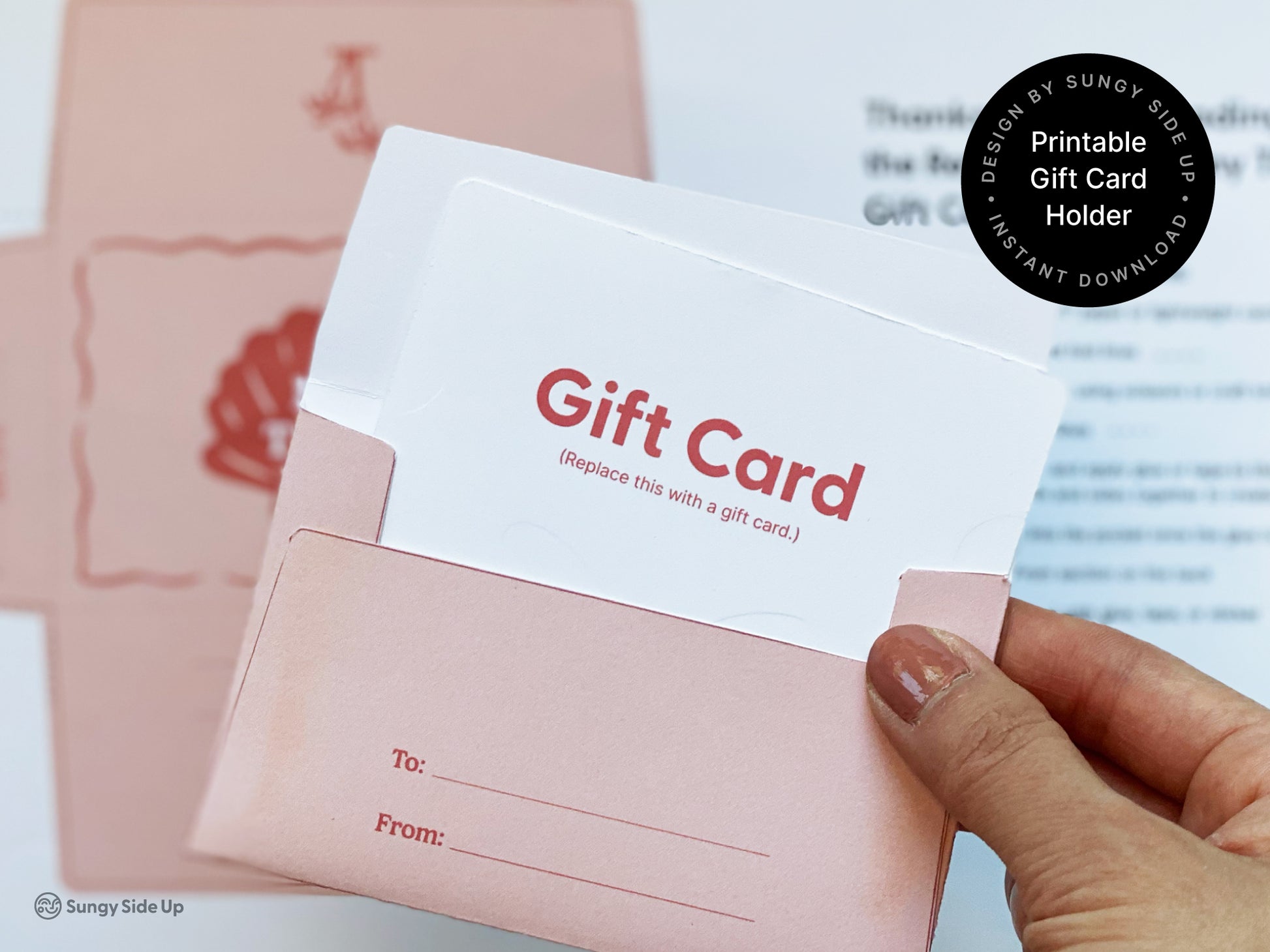 Close-up of a hand holding a pink printable retro-style gift card holder with a mock gift card partially inserted. The holder features a “To” and “From” section for personalization and is part of a DIY assembly project. In the blurred background, printed templates and instructions can be seen, indicating the crafting process. This beach-themed design is ideal for thank-you gifts and is available as an instant digital download for printing on 8.5" x 11" paper at home.