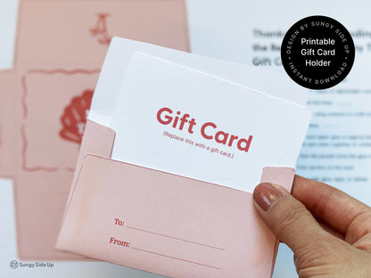 Close-up of a hand holding a pink printable retro-style gift card holder with a mock gift card partially inserted. The holder features a “To” and “From” section for personalization and is part of a DIY assembly project. In the blurred background, printed templates and instructions can be seen, indicating the crafting process. This beach-themed design is ideal for thank-you gifts and is available as an instant digital download for printing on 8.5" x 11" paper at home.