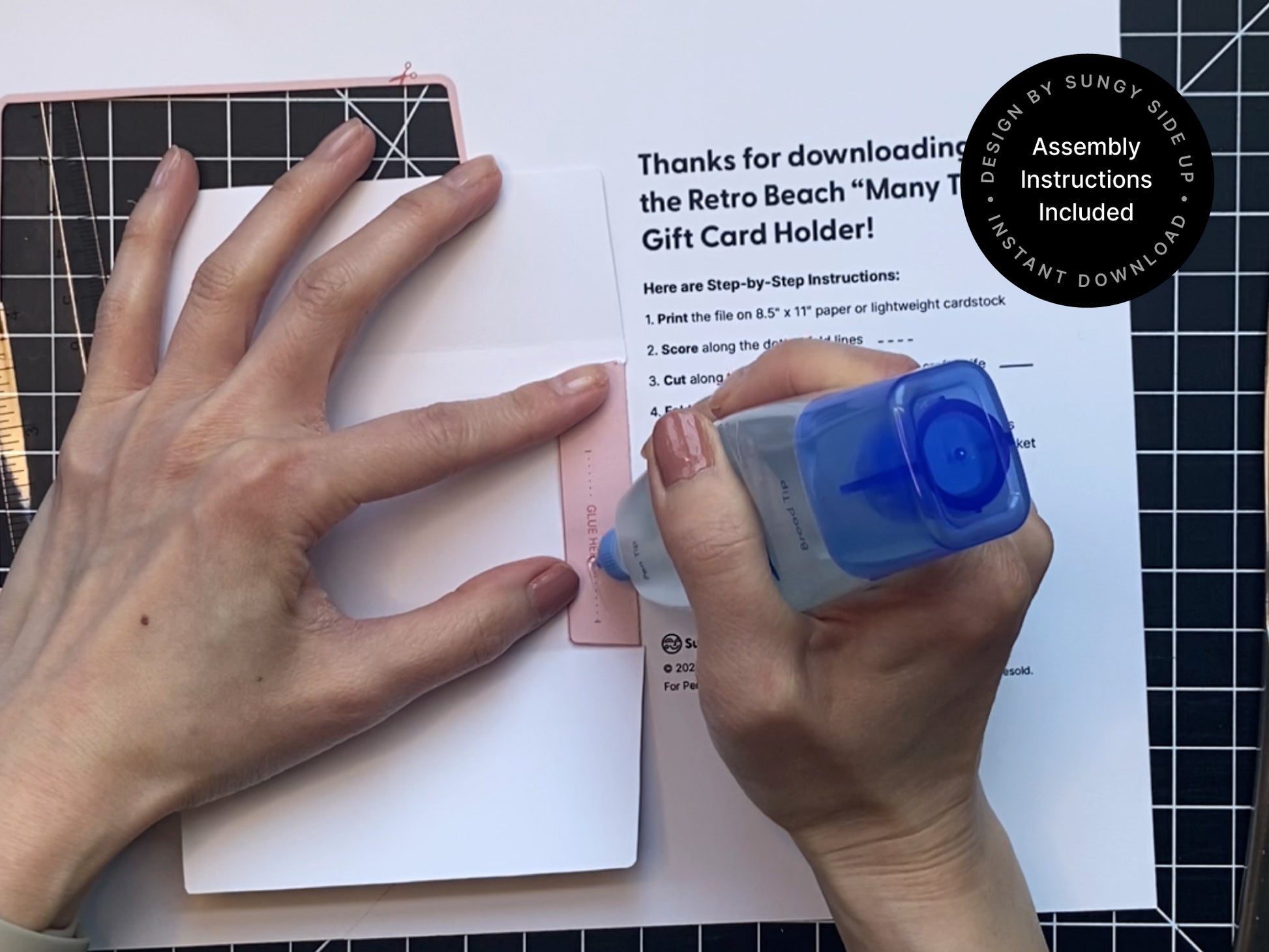 Close-up of hands assembling a printable retro-style gift card holder using glue. The printed template is being adhered along the tab labeled “Glue Here” while a crafting mat and printed step-by-step instructions are visible in the background. The design is made for 8.5" x 11" paper or cardstock and includes a beach-themed pink and red seashell motif. This DIY project is part of an instant digital download that comes with clear assembly instructions for easy at-home crafting.