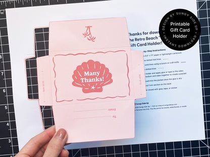 Hand holding an unassembled retro-style printable gift card holder in pink with a red seashell design and “Many Thanks!” on the front. The template is printed on 8.5" x 11" paper and includes glue tabs and “To” and “From” fields. A crafting mat and printed step-by-step instructions are visible in the background. This DIY project is perfect for thank-you gifts, beach-themed events, or special occasions and is available as an instant digital download from Sungy Side Up.