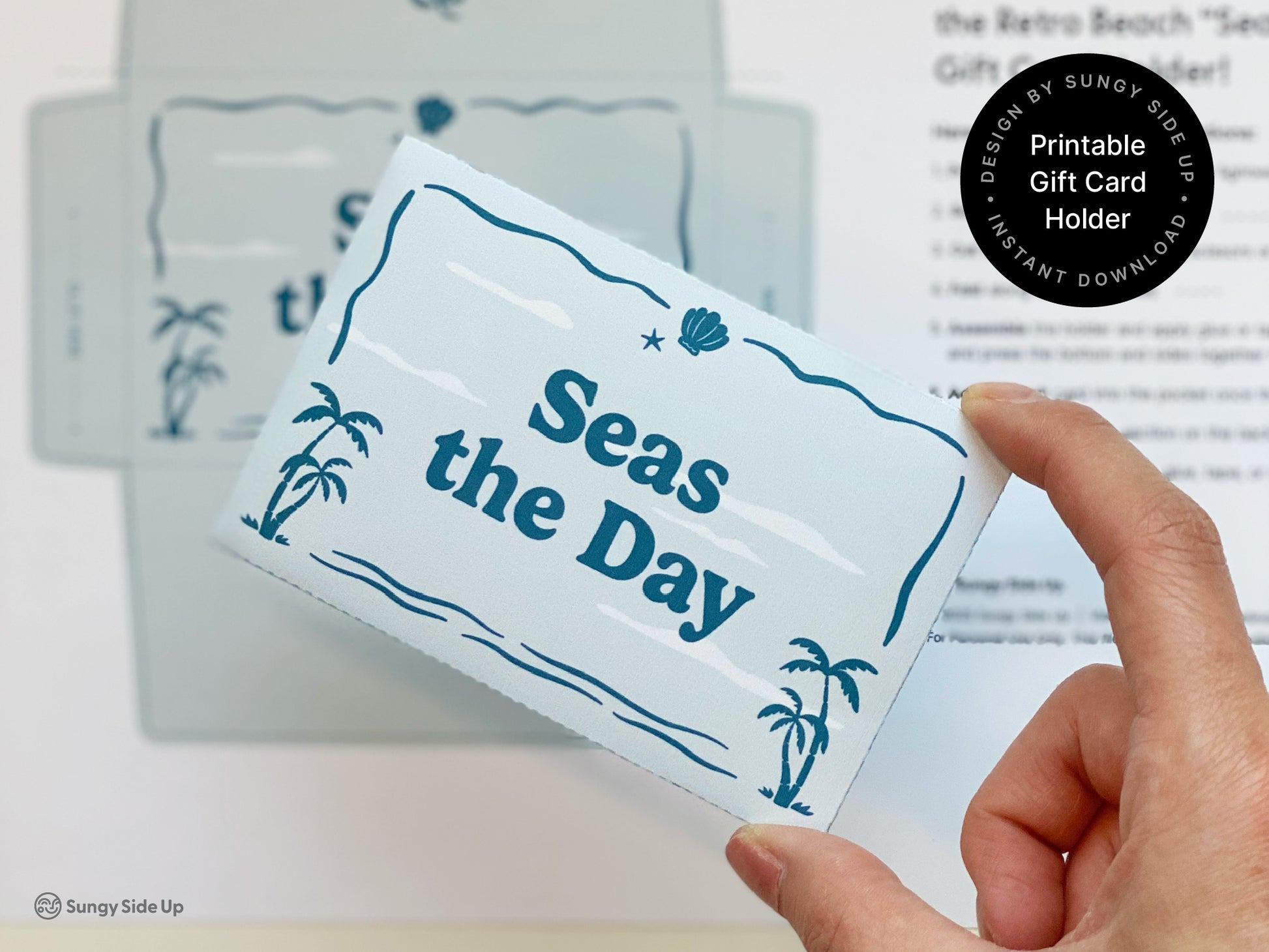 Hand holding an assembled printable gift card holder with a retro beach theme featuring palm trees and ocean inspired details in blue tones. The background shows the printed template and instructions slightly blurred. This DIY project is a digital download designed for easy at home assembly. Ideal for summer gifting birthdays or thank you cards. The design is fun and coastal and perfect for adding a creative handmade touch to a gift card presentation.