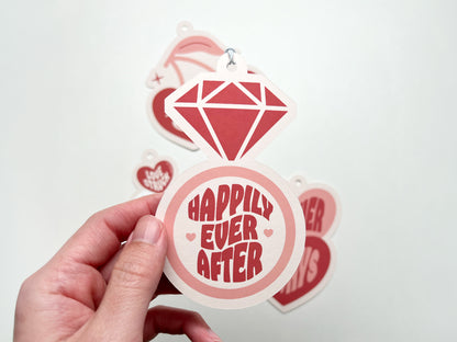 A close-up of a retro-style wedding gift tag in the shape of a diamond ring. The tag features a bold red diamond design and the phrase Happily Ever After in playful retro typography. A hand holds the tag with other tags visible in the background.