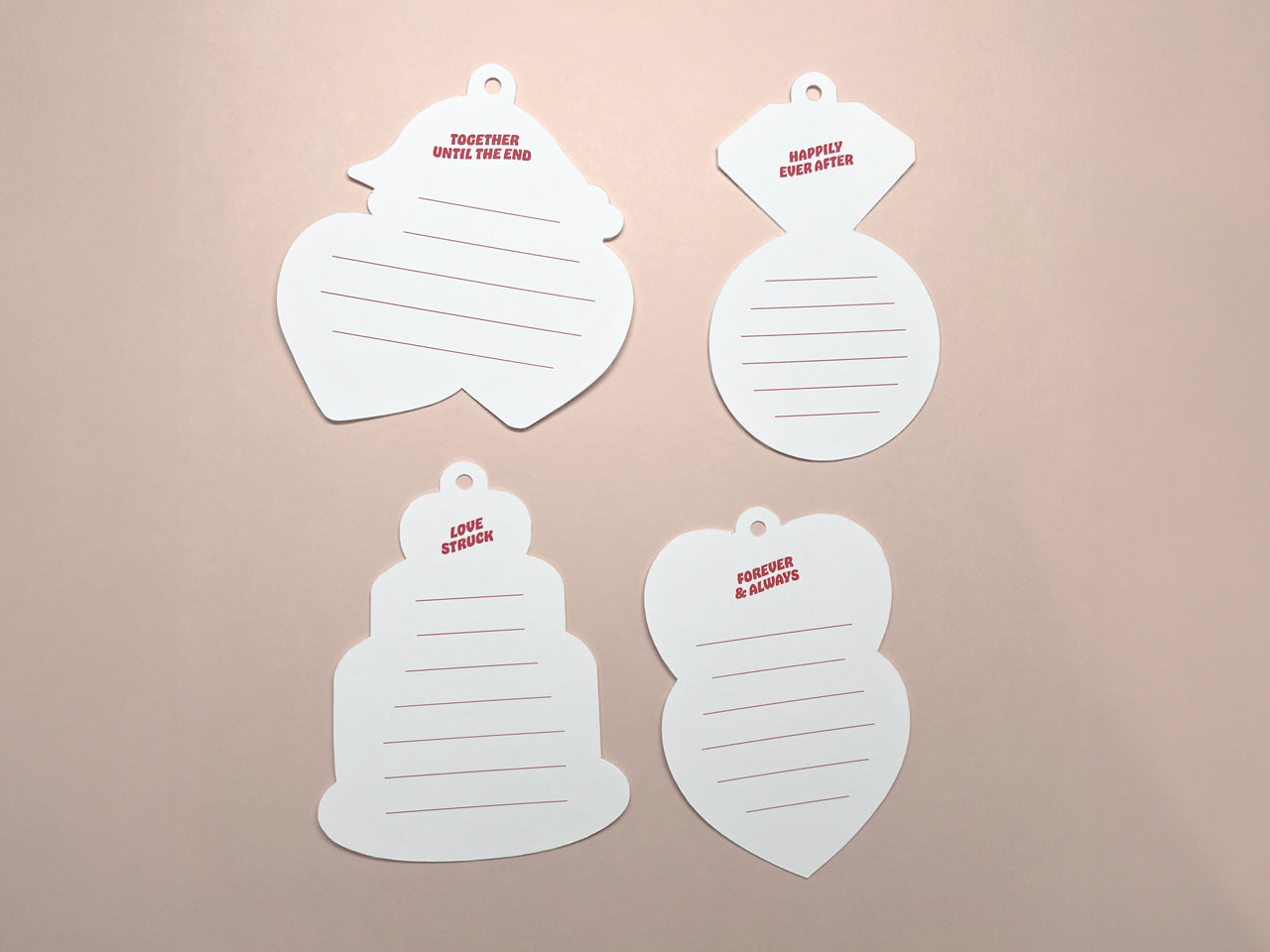 Four retro-style gift tags displayed on a soft pink background, showcasing the blank backs of the tags. Each tag has red lines for writing and small phrases at the top. The tags are shaped like a heart, diamond ring, wedding cake, and stacked hearts.