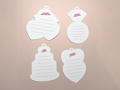 Four retro-style gift tags displayed on a soft pink background, showcasing the blank backs of the tags. Each tag has red lines for writing and small phrases at the top. The tags are shaped like a heart, diamond ring, wedding cake, and stacked hearts.
