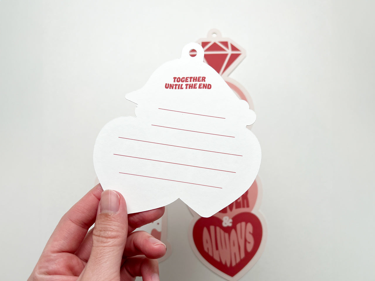 A close-up of the back of a heart-shaped retro-style gift tag with the phrase Together Until the End printed at the top in red. The tag has pre-punched holes for attachment and horizontal red lines for writing.