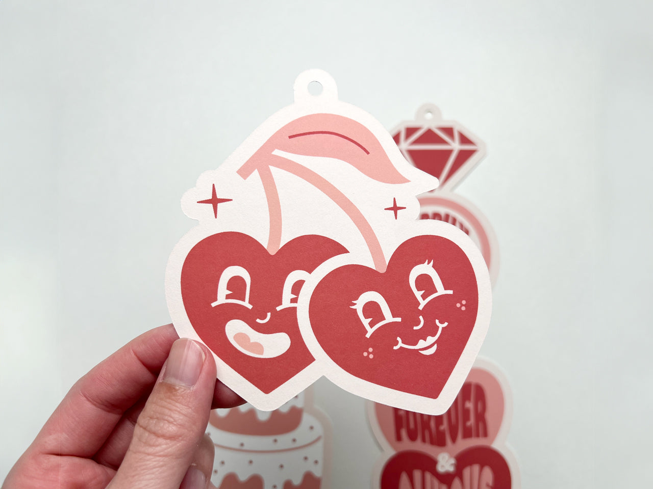 A close-up of a retro-style wedding gift tag shaped like a pair of cherries with heart-shaped faces. One cherry has a wide, happy smile, while the other has a sweet grin. The design uses pink and red hues.