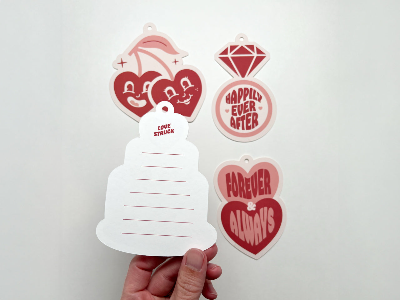 Close-up of the back of a retro-style wedding gift tag shaped like a wedding cake. The tag features red horizontal lines for writing and the phrase Love Struck in bold red text at the top. Other retro-themed tags are displayed in the background.