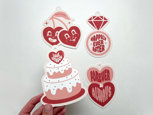 Four retro-style wedding gift tags in pink and red hues. Each tag features a unique design: a pair of cherries with hearts for faces, diamond ring, wedding cake, and two stacked hearts. Each tag has a pre-punched hole at the top for easy attachment.