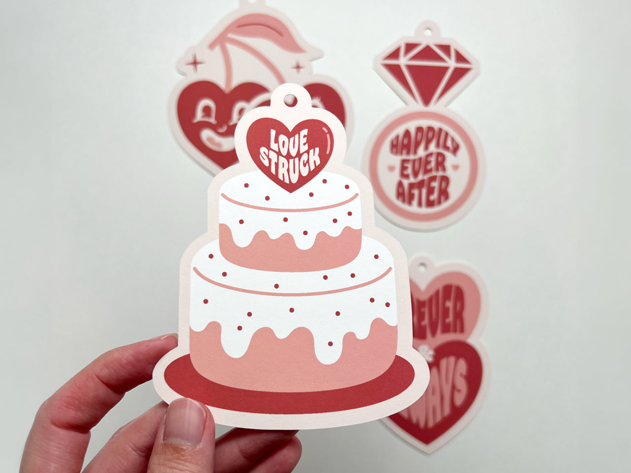A close-up of a retro-style wedding gift tag shaped like a two-tiered wedding cake. The cake has white frosting with red polka dots and a pink heart topper that reads Love Struck in bold white text.
