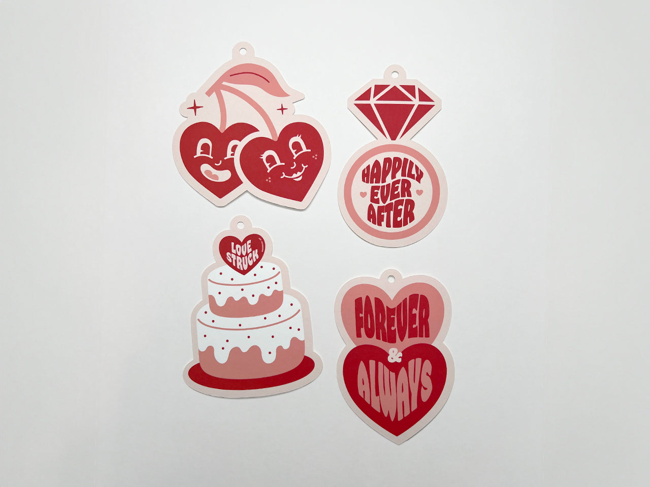 A set of four retro-style wedding gift tags featuring playful designs and bold text in pink and red hues: a pair of smiling cherries with heart-shaped faces, a diamond ring, a wedding cake, and two stacked hearts. Each tag has pre-punched holes.