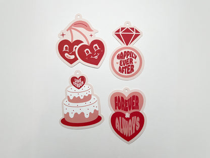 A set of four retro-style wedding gift tags featuring playful designs and bold text in pink and red hues: a pair of smiling cherries with heart-shaped faces, a diamond ring, a wedding cake, and two stacked hearts. Each tag has pre-punched holes.