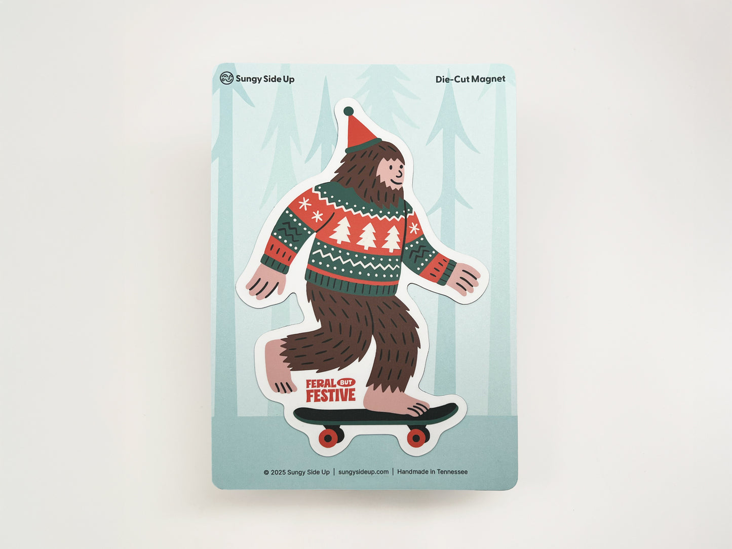 Die-cut Christmas Sasquatch magnet featuring Bigfoot on a skateboard wearing a festive sweater and Santa hat, displayed on Sungy Side Up backing card.