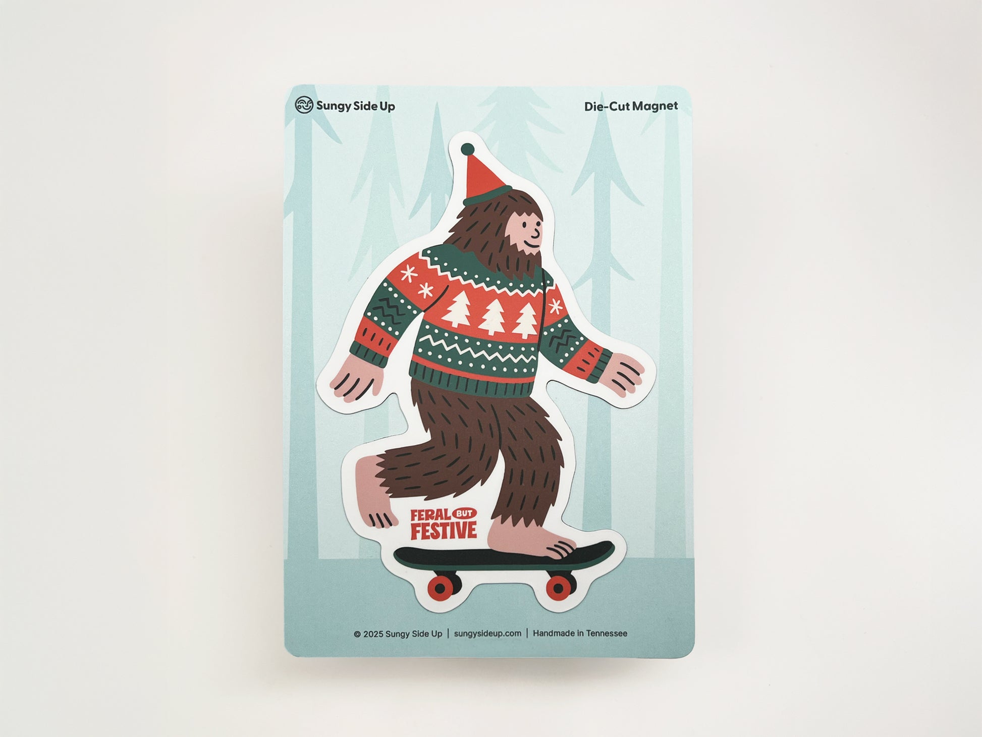 Die-cut Christmas Sasquatch magnet featuring Bigfoot on a skateboard wearing a festive sweater and Santa hat, displayed on Sungy Side Up backing card.