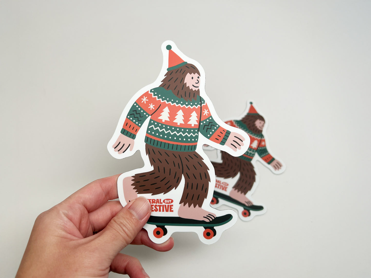 Close-up of Feral but Festive Bigfoot magnet held in hand, showing detailed die-cut shape and colorful Christmas sweater design.