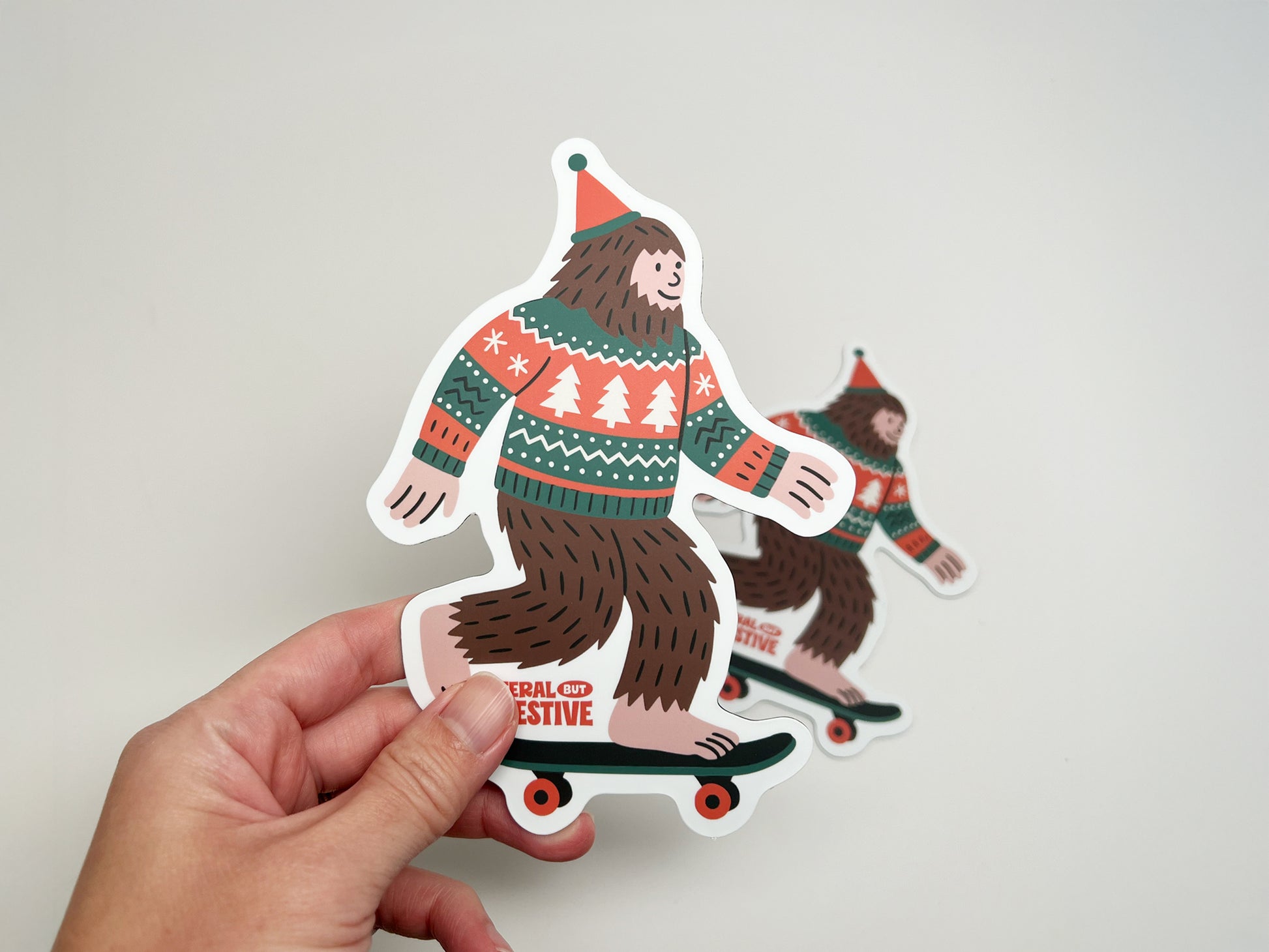 Close-up of Feral but Festive Bigfoot magnet held in hand, showing detailed die-cut shape and colorful Christmas sweater design.