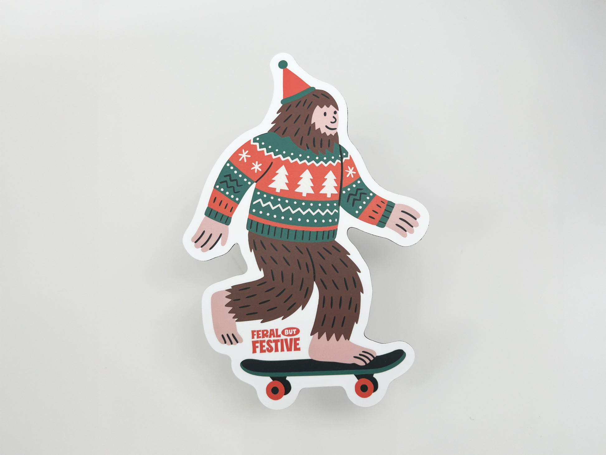 Flat lay of die-cut Sasquatch magnet wearing Christmas sweater and hat, riding skateboard with “Feral but Festive” text, holiday decor gift idea.