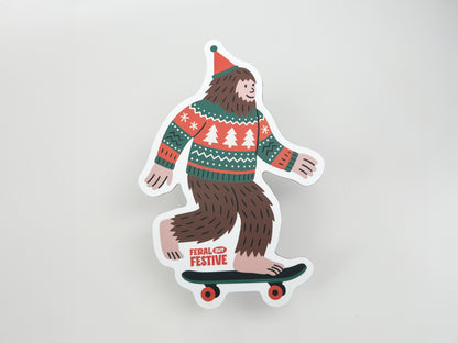 Flat lay of die-cut Sasquatch magnet wearing Christmas sweater and hat, riding skateboard with “Feral but Festive” text, holiday decor gift idea.