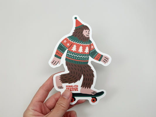 Person holding festive Sasquatch magnet up close, showing Bigfoot on skateboard in red and green holiday sweater, handmade Christmas magnet.