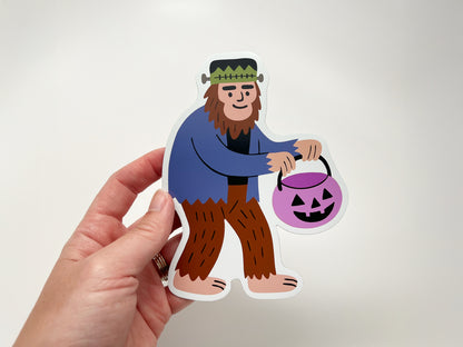 Hand holding a die-cut Halloween magnet featuring a quirky Sasquatch dressed as Frankenstein and carrying a purple jack-o’-lantern candy bucket. Fun seasonal fridge magnet, perfect for cryptid fans, spooky décor lovers, and unique Halloween gifts or collectible novelty magnets.