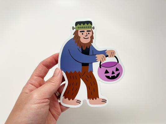 Hand holding a die-cut Halloween magnet featuring a quirky Sasquatch dressed as Frankenstein and carrying a purple jack-o’-lantern candy bucket. Fun seasonal fridge magnet, perfect for cryptid fans, spooky décor lovers, and unique Halloween gifts or collectible novelty magnets.