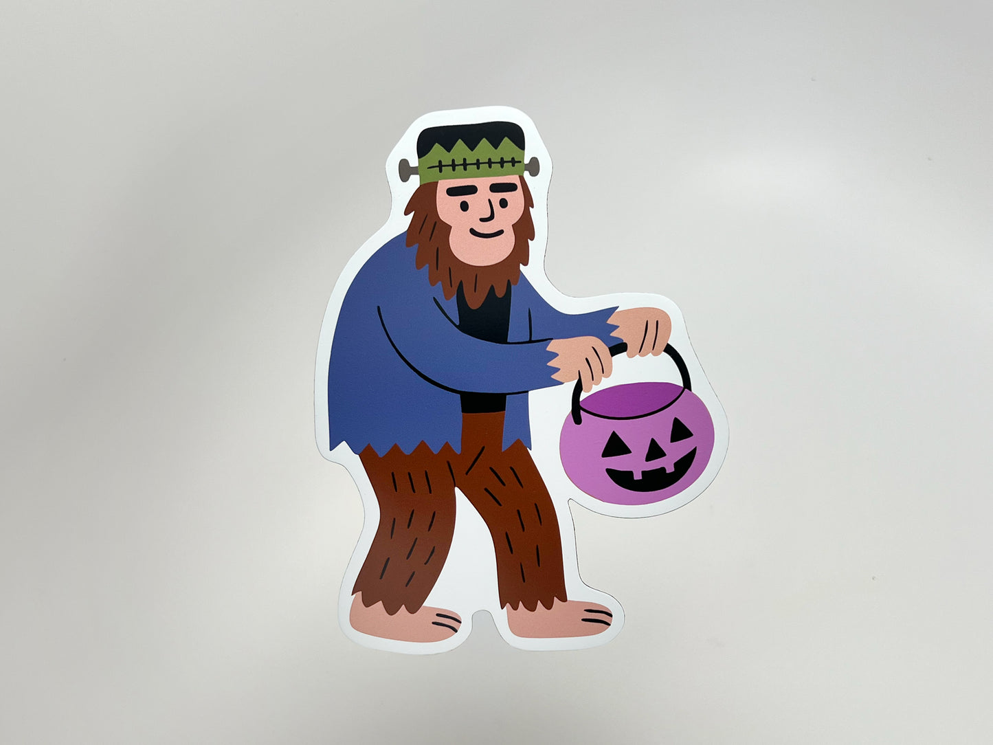 Die-cut Halloween magnet featuring a friendly Sasquatch dressed as Frankenstein, holding a purple jack-o’-lantern candy bucket. Fun and quirky seasonal décor for fridges, lockers, or offices. Unique cryptid Halloween gift idea, perfect for spooky season lovers and collectors of offbeat magnets.