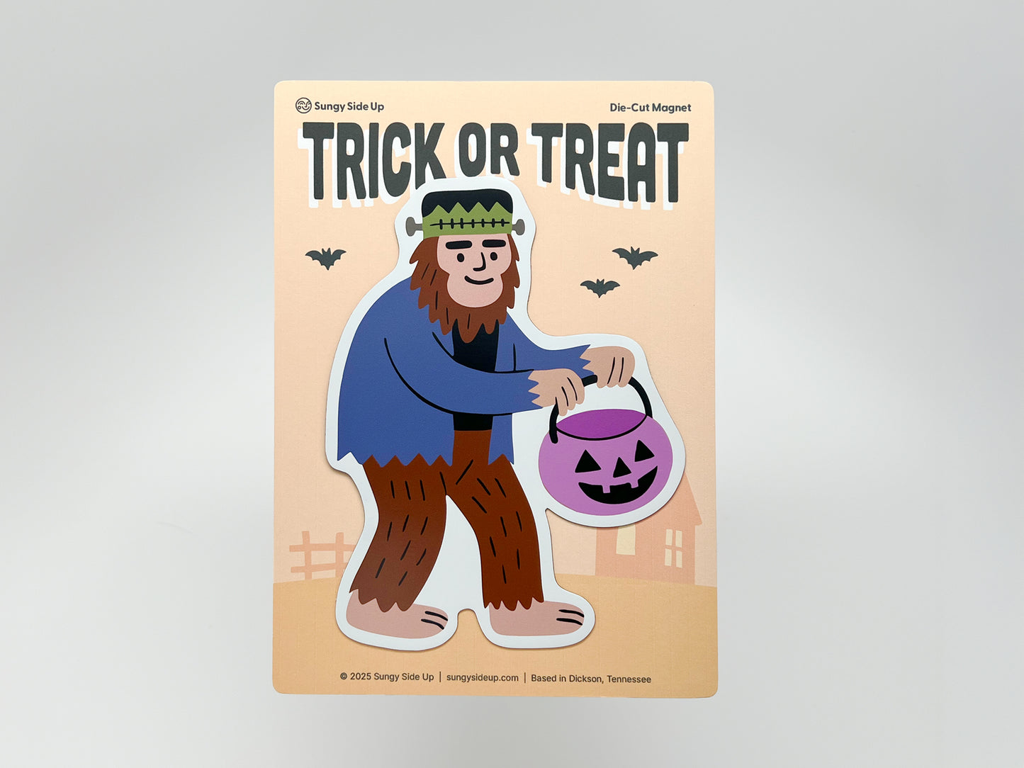 Halloween die-cut magnet featuring a quirky Sasquatch dressed in a Frankenstein costume holding a purple pumpkin candy bucket. Fun cryptid Halloween décor, perfect for fridges, lockers, or offices. Unique seasonal magnet gift for spooky season fans and lovers of quirky home accessories.