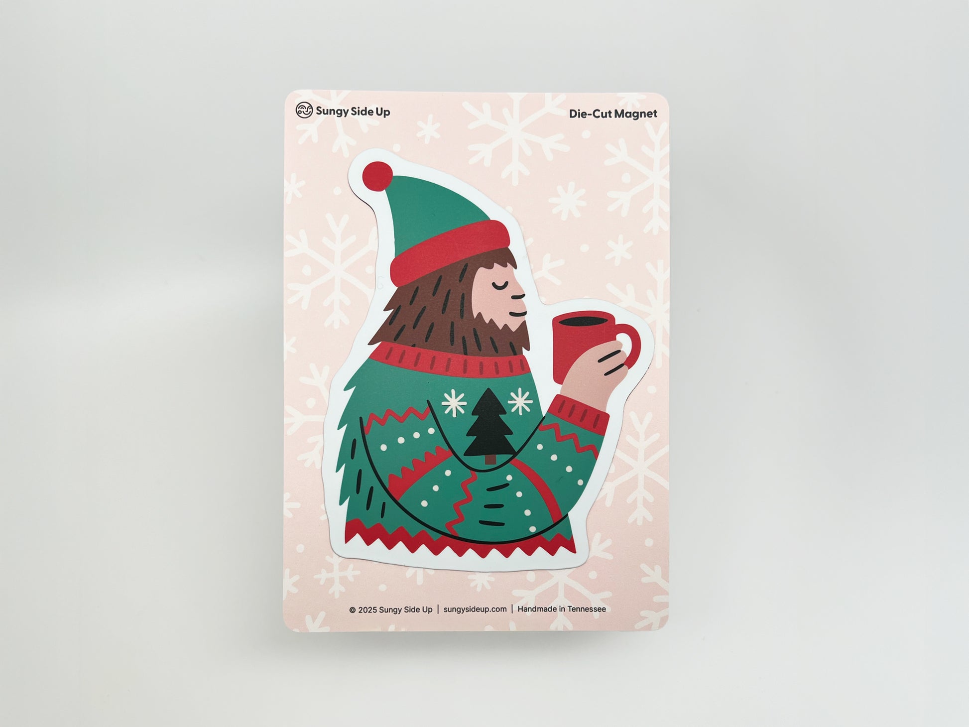 Illustration of a cheerful Sasquatch wearing a green and red winter sweater and matching hat, holding a red mug of hot cocoa with a peaceful smile. The die-cut magnet is displayed on a pink backing card decorated with white snowflakes, branded with “Sungy Side Up” and labeled “Die-Cut Magnet” at the top.