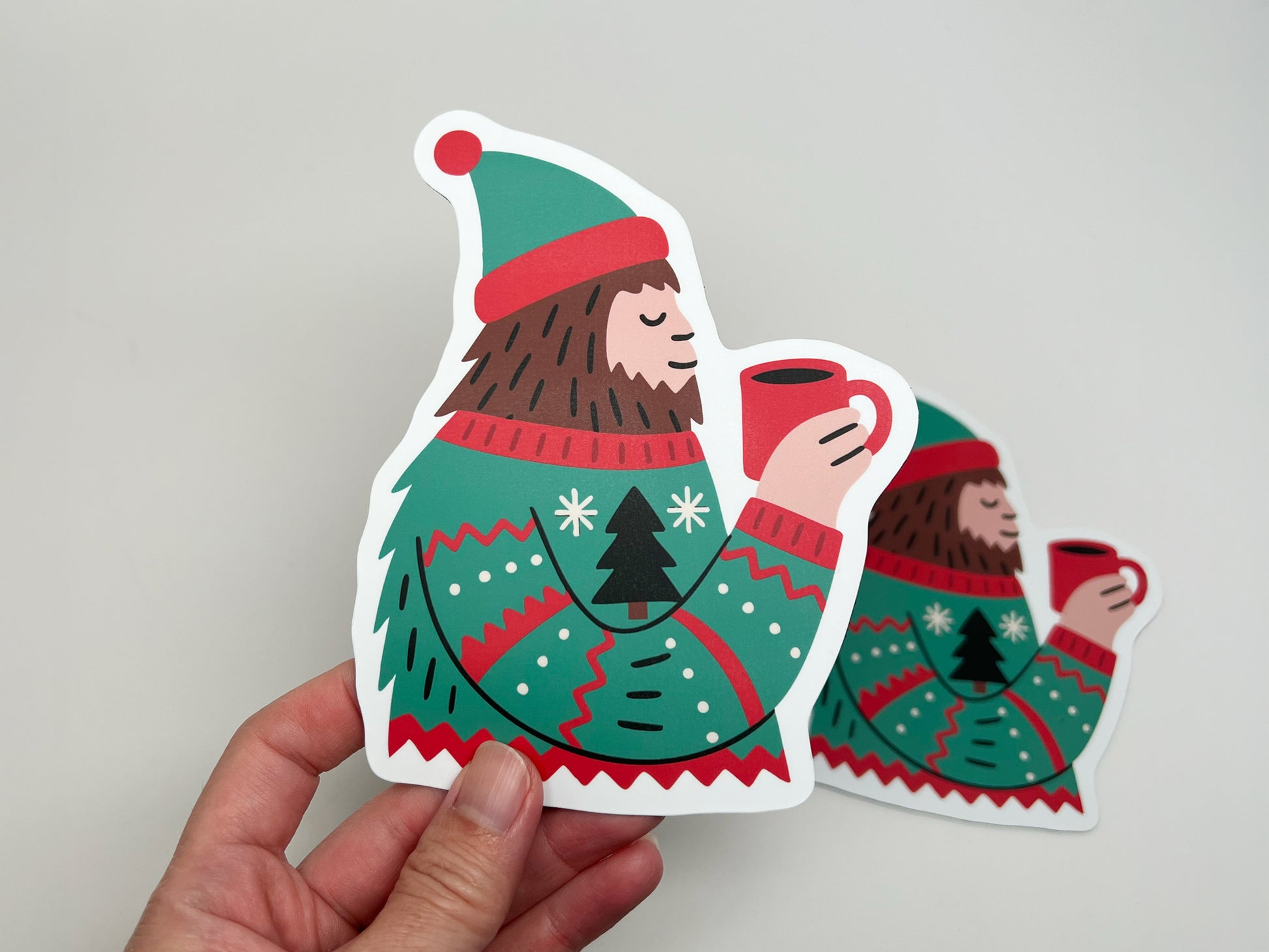 A hand holding a die-cut magnet shaped like a Sasquatch wearing a festive green and red sweater with a Christmas tree design, holding a red mug of hot cocoa. Another identical magnet is slightly blurred in the background against a plain light surface, highlighting the product’s colorful details and glossy finish.