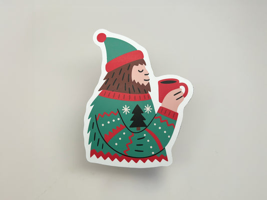 Die-cut magnet shaped like a Sasquatch wearing a green and red winter sweater with a Christmas tree design and matching hat, holding a red mug of hot cocoa. The magnet is photographed against a plain light gray background, showing its vibrant colors and clean cut edges.