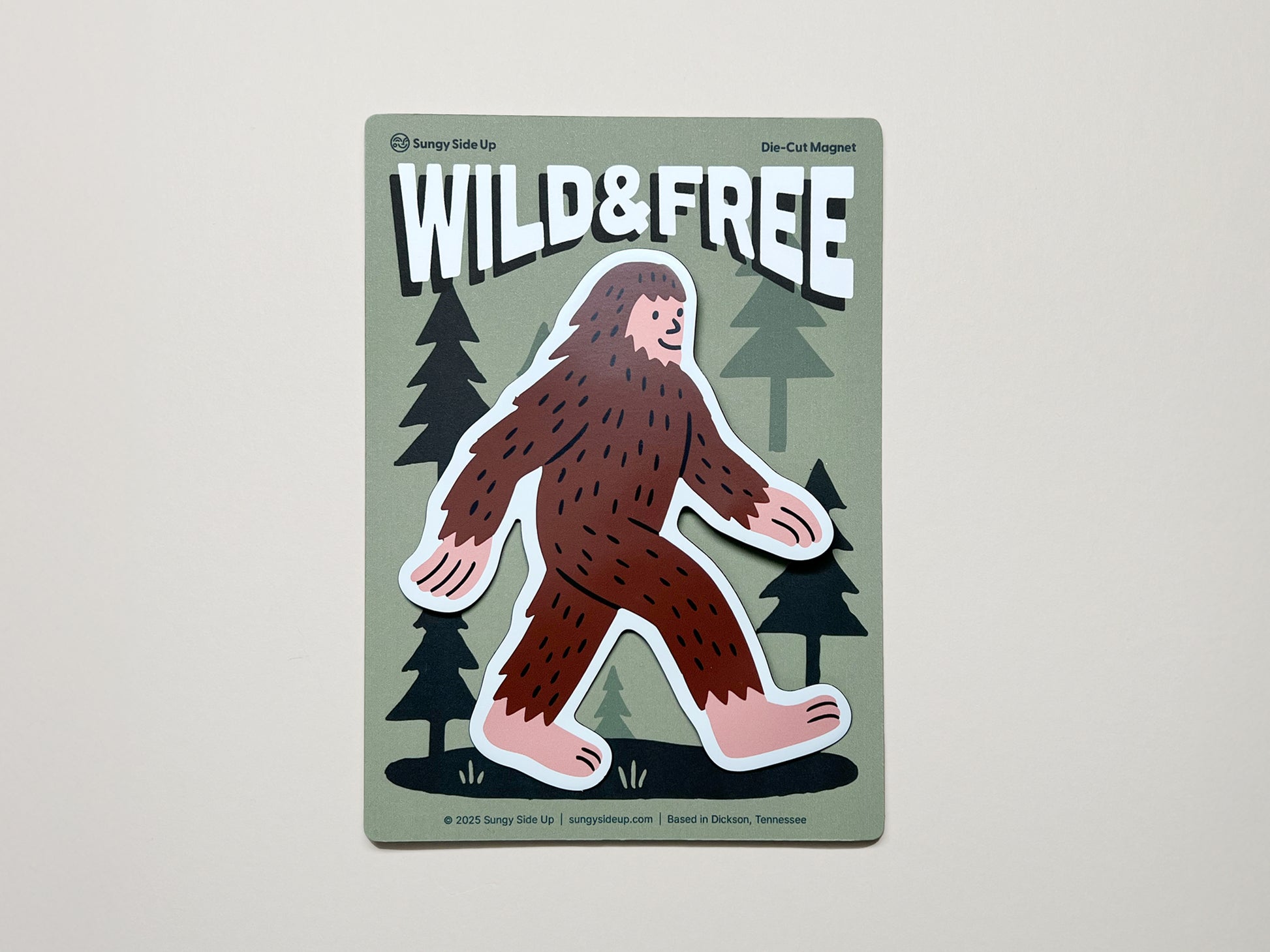 Handmade die-cut magnet featuring a cute brown Sasquatch walking through a forest with the phrase "WILD & FREE" above; quirky Bigfoot fridge magnet by Sungy Side Up, perfect for nature lovers, cryptid fans, and whimsical gift giving.