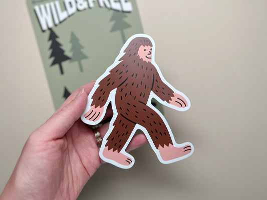 Hand holding a large die-cut Bigfoot magnet with brown fur and pink feet; quirky Sasquatch refrigerator magnet from Sungy Side Up, perfect gift for cryptid lovers, outdoorsy friends, and fun stocking stuffers.