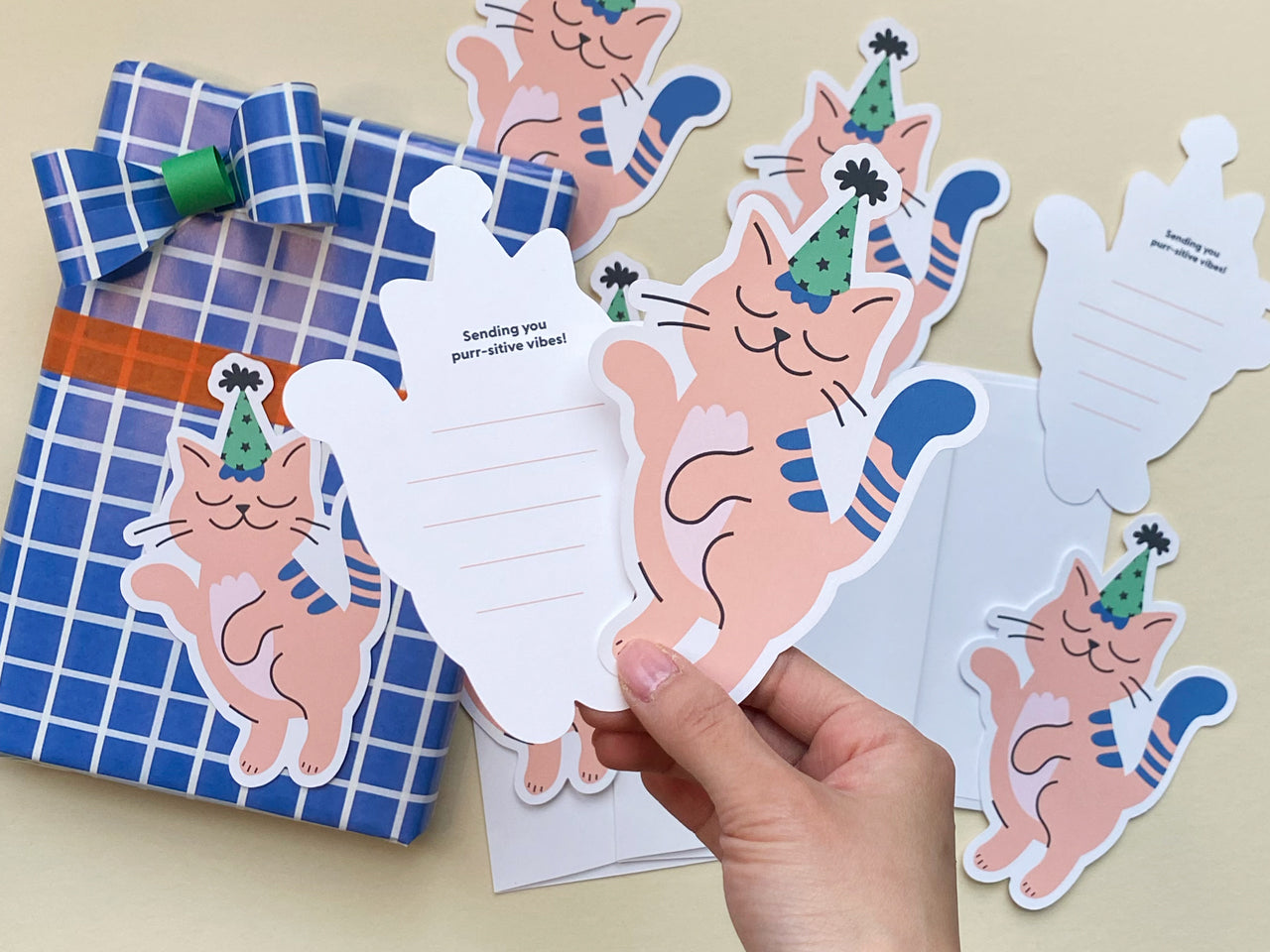 A hand holds a die-cut flat note card featuring a pink cat wearing a green and black party hat with a playful dancing pose. The back of the card has lined spaces with the words Sending you purr-sitive vibes! written at the top.