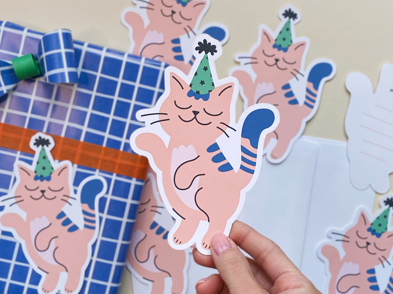 A hand holds a die-cut note card featuring a pink cat wearing a green and black party hat and looking joyful. The front of the card is visible, showing the full design. In the background, there is a gift wrapped in blue grid-patterned paper.