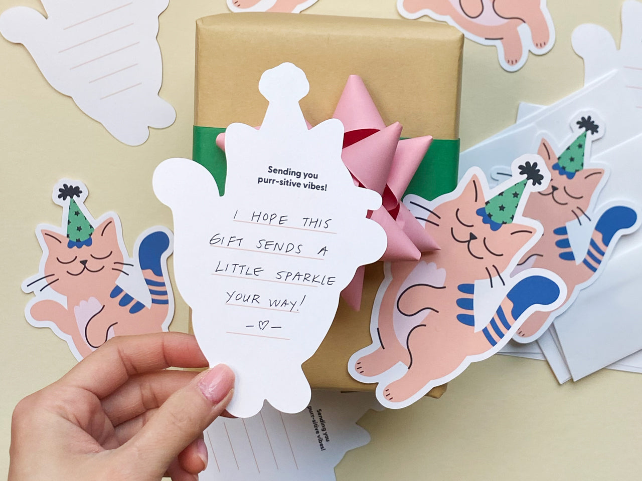 A hand holds a die-cut note card showing handwritten text on the back. The card also has the pre-printed text Sending you purr-sitive vibes! at the top. The scene includes a small wrapped gift with a pink bow, more cat note cards, and envelopes.